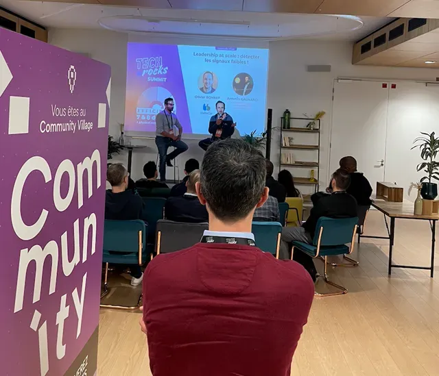 Session au Community Village du Tech.Rocks Summit : deux intervenants sur scène devant l’écran, public assis, kakémono « Vous êtes au Community Village » au premier plan